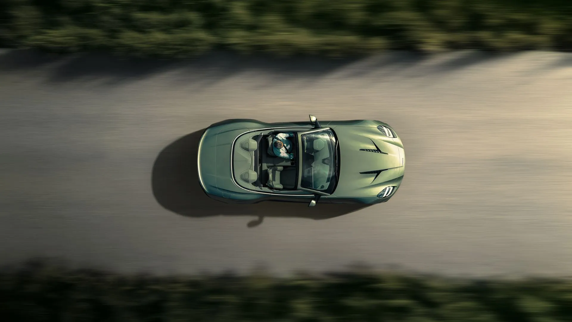 Aston Martin Vanquish Volante mui trần màu xanh lá cây nhìn từ trên cao, đang lướt trên con đường quê uốn lượn, thể hiện sự sang trọng và hiệu suất của dòng xe V12.