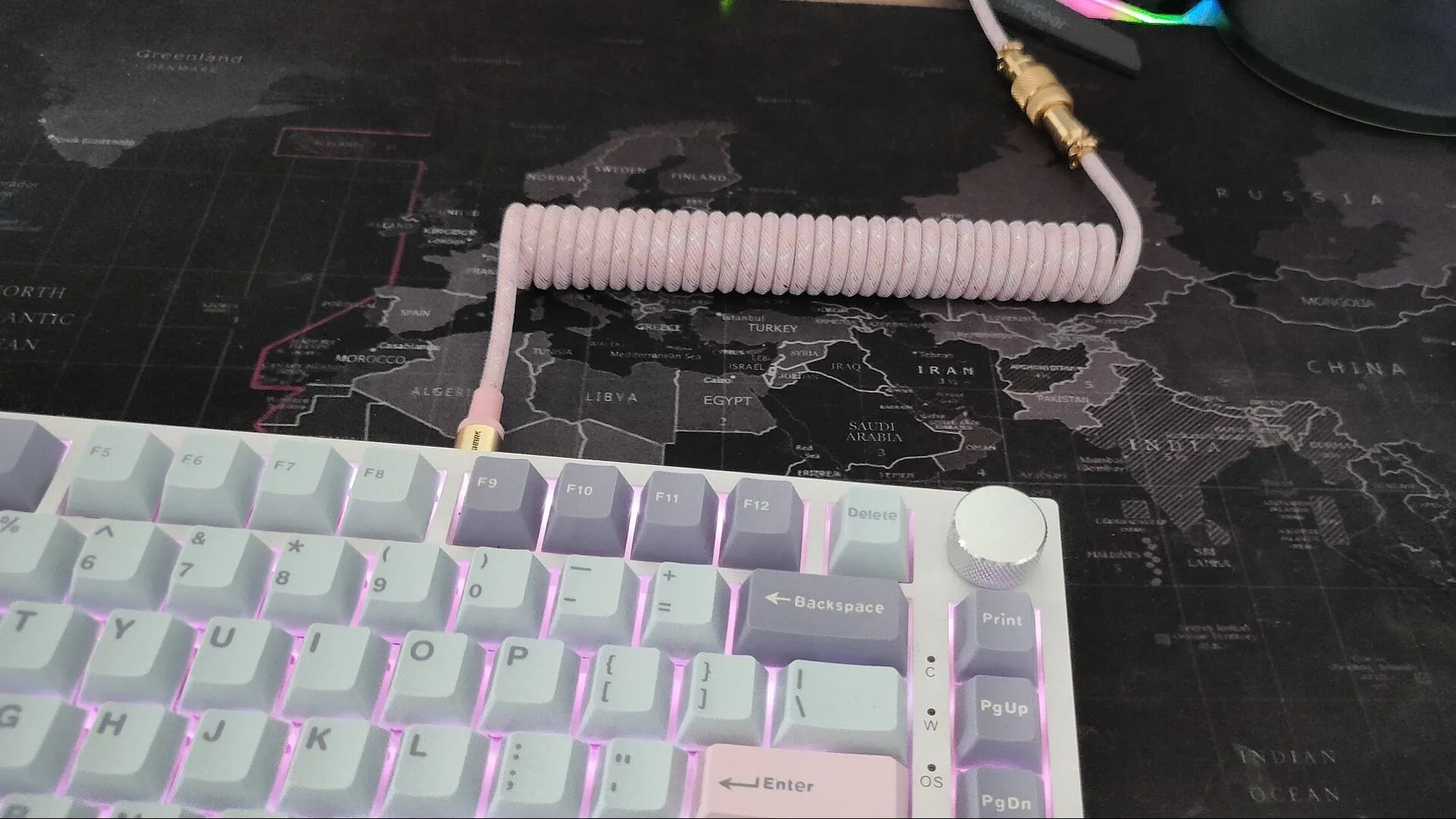 Dây cáp bàn phím xoắn màu hồng cắm vào bàn phím cơ màu trắng với keycaps xanh và tím, tạo điểm nhấn độc đáo.