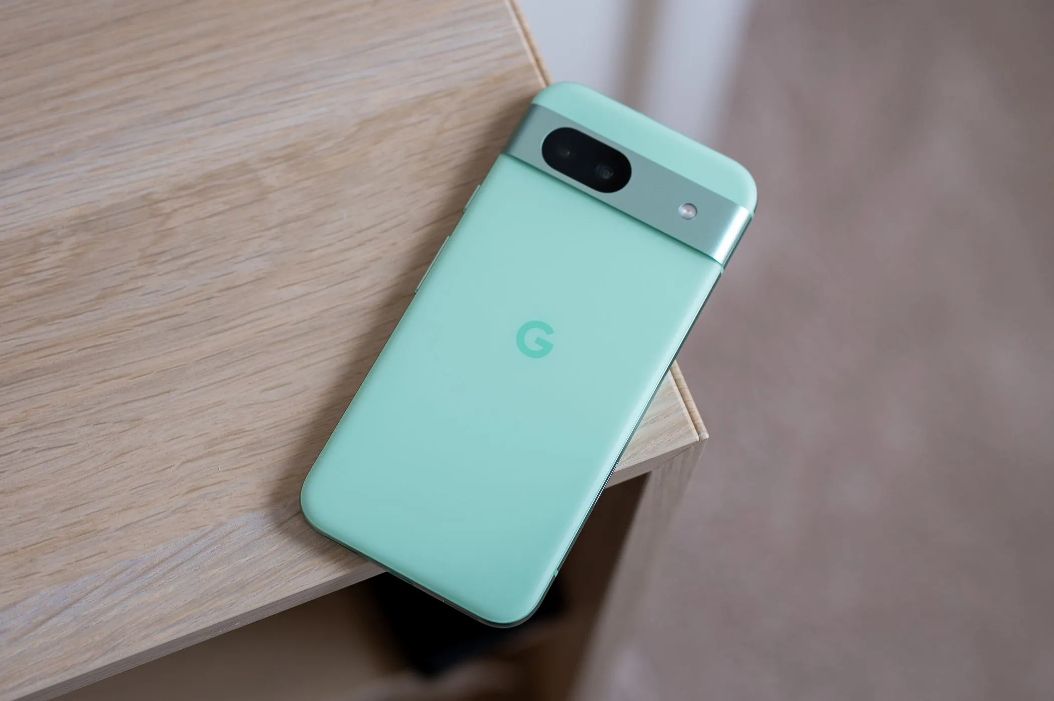 Điện thoại Google Pixel 8a màu xanh Aloe, đặt nghiêng trên mép bàn, cho thấy thiết kế dải camera ngang đặc trưng và mặt lưng cong nhẹ.