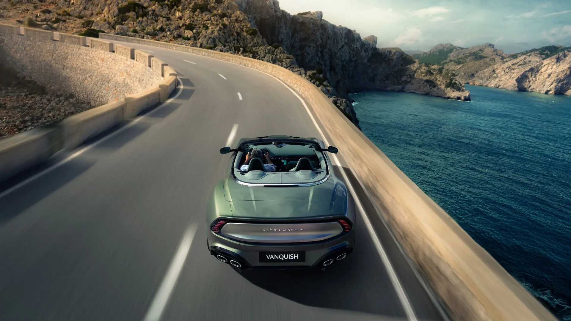 Góc nhìn từ phía sau của Aston Martin Vanquish Volante mui trần màu đen, đang di chuyển trên con đường quanh co bên bờ hồ, khoe thiết kế thể thao và đèn hậu LED đặc trưng.