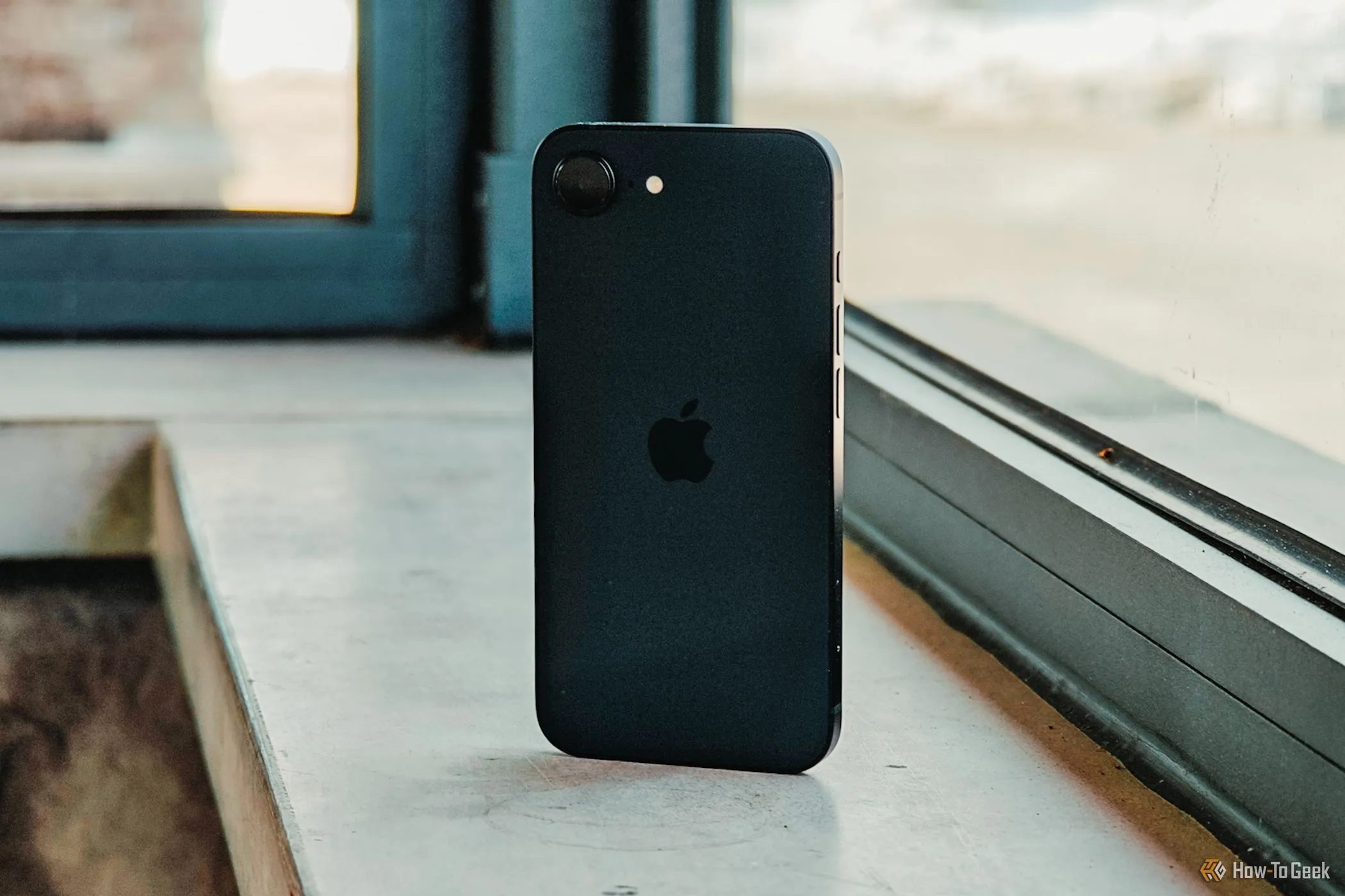 iPhone 16e màu trắng được cầm trong tay, phần mặt lưng nhìn nghiêng dựa vào cửa sổ, nổi bật logo Apple