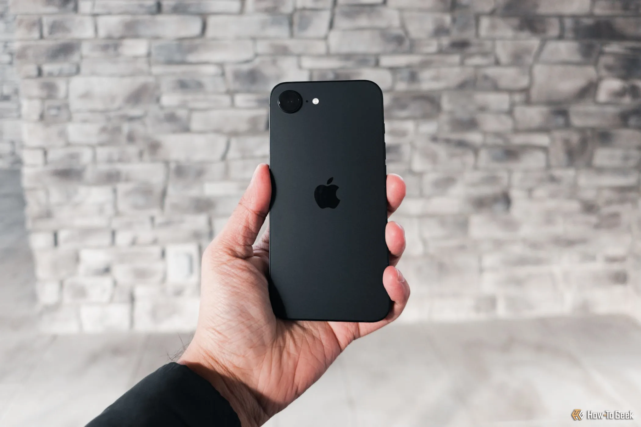 Người dùng cầm iPhone 16e màu trắng trên tay, nổi bật ở giữa, đứng trước bức tường xám, thể hiện tính di động và sự tiện lợi