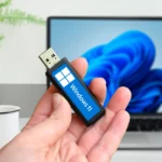 Người dùng cầm USB cài đặt Windows 11, phía sau là laptop đang hiển thị giao diện hệ điều hành.